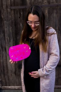Mama mit braunen Haaren und Brille, eine Hand auf dem Bauch, in der anderen ein Schild mit "hallo baby" - zweite Schwangerschaft verkünden