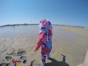 kleines Mädchen am Strand, von hinten zu sehen, steht und zeigt in Richtung Meer, hat einen bunten UV-Anzug an und Sandalen von Crocs, daneben liegt Sandspielzeug im Sand