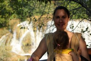 Mama mit Klinkind in der Trage vor dem Bauch - im Hintergrund die Wasserfälle im Nationalpark Krka in Kroatien