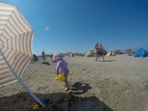kleines zwei jähriges Mädchen am Strand mit Sonnenhut und UV-Shirt hat eine gelbe Gießkanne in der Hand, links ein orange weuß gestreifter Sonnenschirm