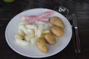 Abendsessen auf einem weißen Teller angerichtet: weißer Spargel geschnitten mit ungeschälten Kartoffeln, heller Soße und Röllchen von gekochtem Schinken