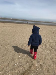 Kind von hinten mit roten Gummistiefeln und blauer Fleecejacke mit Öhrchen läuft durch Sand am Strand in Südholland