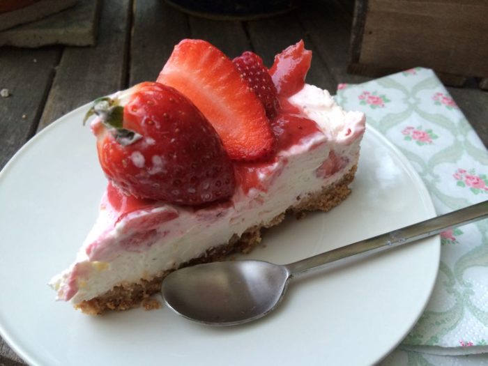 Ein Stück Kuchen mit Frischkäsecreme und Erdbeeren auf weißem Teller mit silbernem Kaffelöffel: mit Erdbeeren garnierter No bake Kuchen